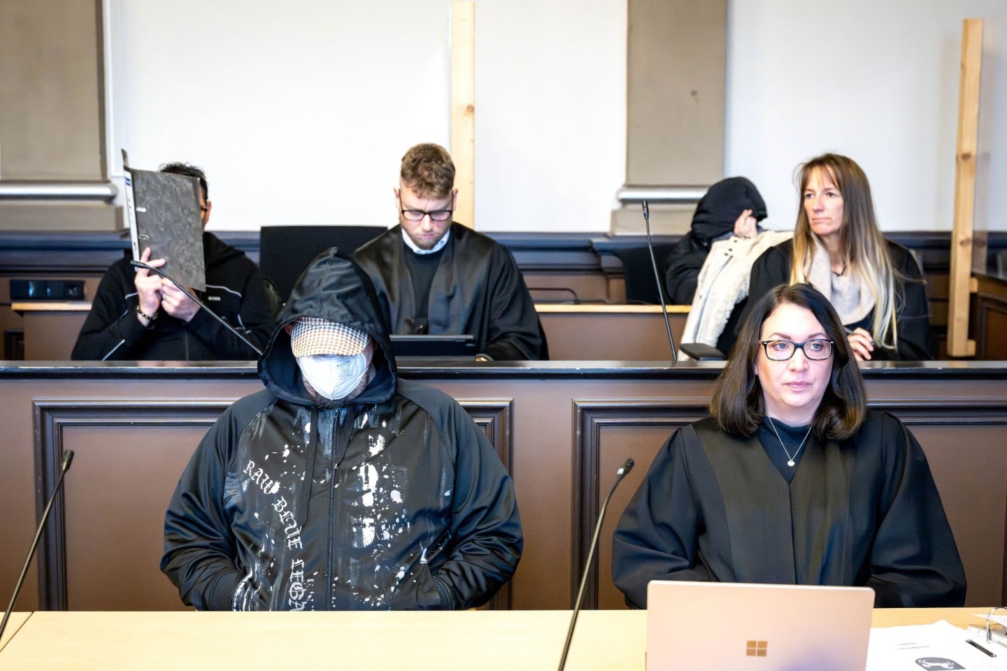Die drei Angeklagten (l, vorne l sowie hinten r) sitzen vor Prozessbeginn mit ihren Anwältinnen und Anwälten im Gerichtssaal.