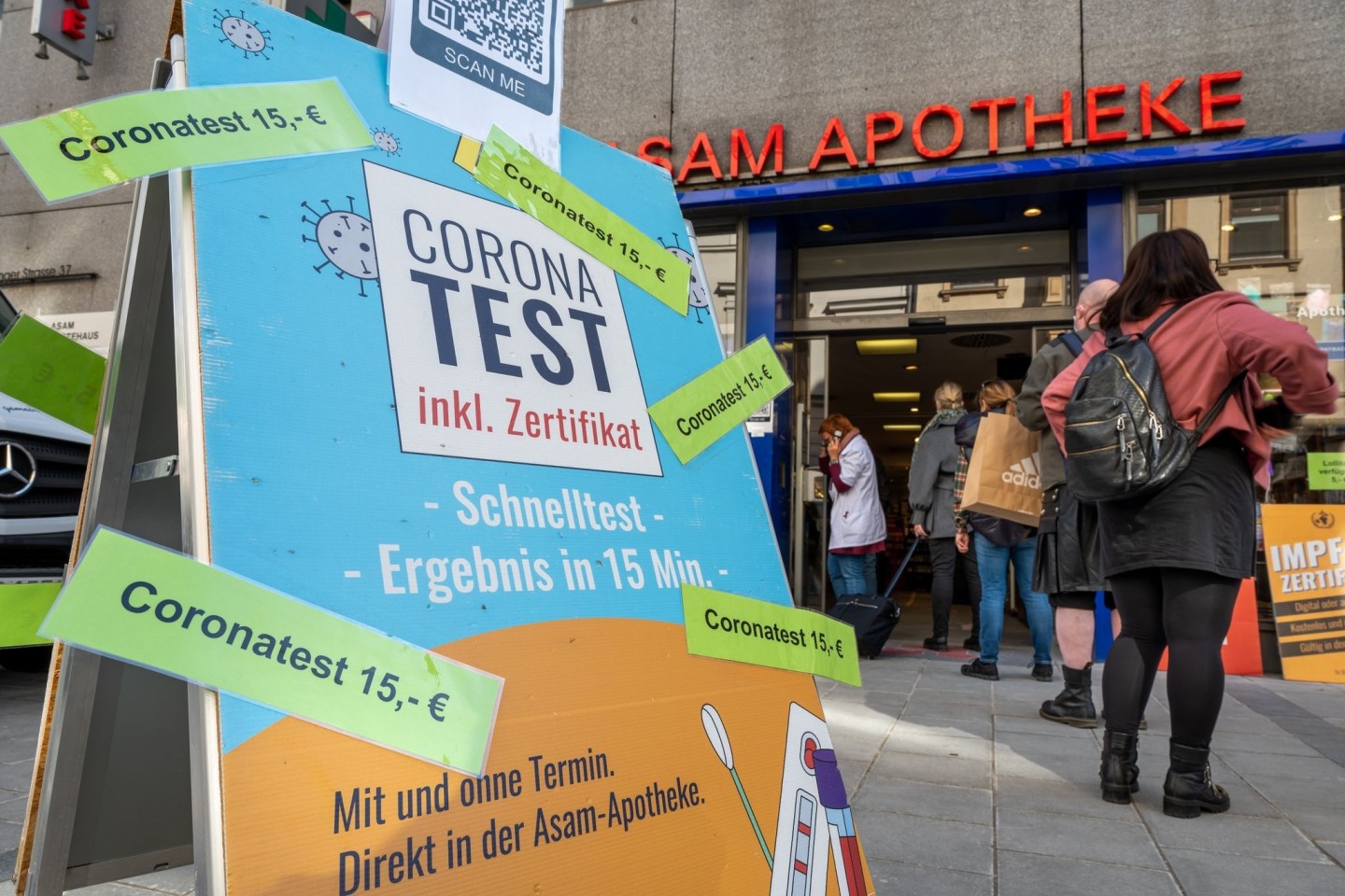 Passanten stehen in der Münchner Innenstadt vor einer Apotheke, die Coronatest für 15 Euro anbietet.